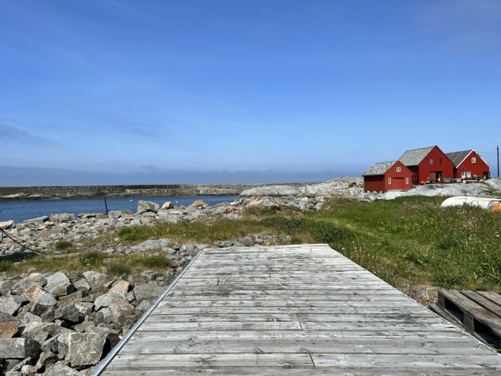 Gammel brygge på Grip med blå himmel og blått hav og tre røde hus mot grønt gress